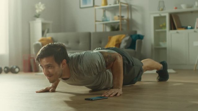 Athletic Fit Man in T-shirt and Shorts is Doing Push Up Exercises While Using a Stopwatch on His Phone. He is Training at Home in His Living Room with Minimalistic Interior.