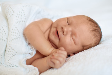 Laughing, smiling  little newborn baby boy sleeping in white blanket, lying on bed.