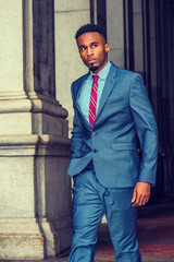 Young African American Businessman with beard traveling, working in New York, wearing dark sky blue suit, white striped shirt, violet red patterned tie, walking by columns on vintage street, looking..