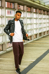 Young Man Casual Fashion in New York. Young African American Guy with beard, wearing black leather jacket, white shirt, black pants, sneakers, standing on walk way with glass wall and wooden floor.