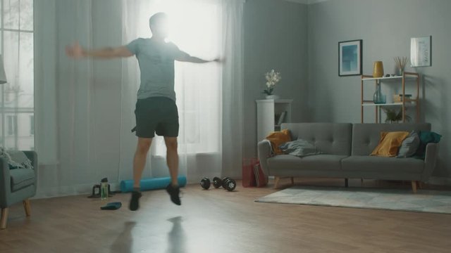 Strong Athletic Fit Man In T-shirt And Shorts Is Doing Jumping Jack Exercises At Home In His Spacious And Bright Apartment With Minimalistic Interior.