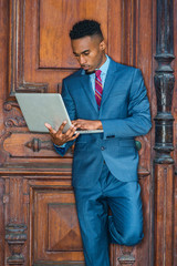 Way to Success. Young African American Businessman with beard working in New York, wearing sky blue suit, violet red patterned tie, standing by brown vintage office door, working on laptop computer..