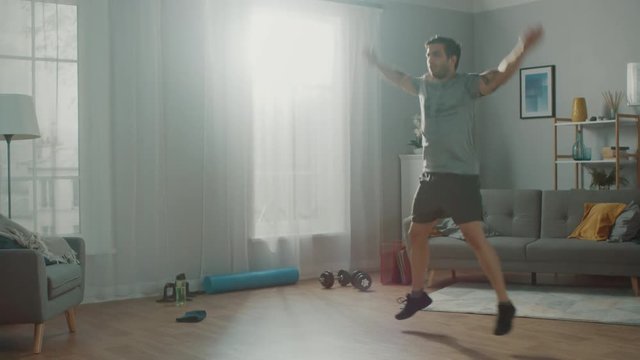 Muscular Athletic Fit Man in T-shirt and Shorts is Doing Jumping Jack Exercises at Home in His Spacious and Bright Apartment with Minimalistic Interior.