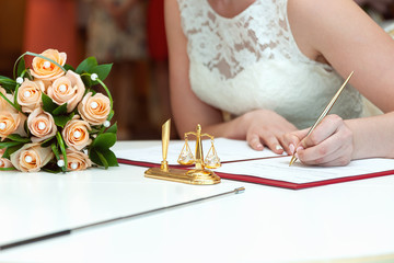 The bride signs the documents during marriage registration. Next to the table is a bridal bouquet of roses. Only hands, close-up.
