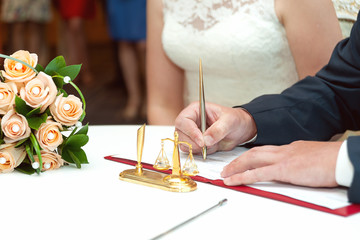 The groom signs the documents during marriage registration. Next to the table is a bridal bouquet of roses. Only hands, close-up.
