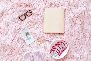 Flatlay of women reading glasses, book, fresh sliced dragon fruit, shoes, accessories on a pink fur background