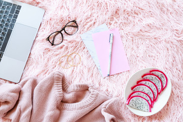 Flatlay of laptop, women knitted sweater, reading glasses, paper, pen, fresh sliced dragon fruit, accessories on a pink fur background