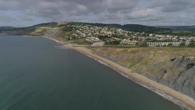 Aerial tracking from sea in towards the village of Charmouth. Amazing Jurassic coast cliffs surround the holiday destination either side. Amazing sunny day.  Vegetation in full bloom