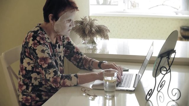 An old woman sits at the table, a cosmetic tissue mask is applied to her face. She uses a computer. Facial care. Plastic surgery and collagen injections. Realistic images with their own imperfections.