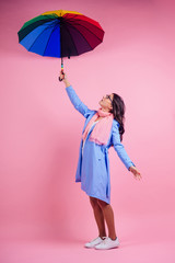 beautiful brunette girl in the studio on a pink background in the style blue raincoat and chiffon scarf with rainbow an umbrella. autumn fashion look seasonal spring sale