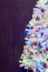 Christmas background. White artificial Christmas tree, decorated with purple Christmas-tree decorations and a garland with lights, on a purple melange background. With free space. 
