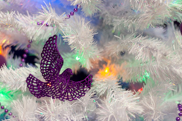 Christmas background. Purple Christmas-tree decoration in the form of a bird against a white artificial Christmas tree, illuminated by lights of garlands. Selective focus, bokeh.
