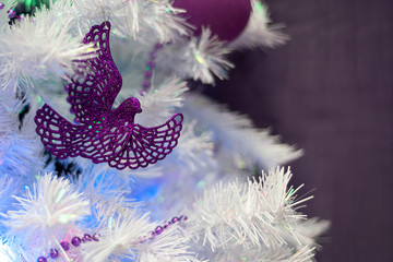 Christmas background. Purple Christmas-tree decoration in the form of a bird against a white artificial Christmas tree, illuminated by lights of garlands. Selective focus, bokeh.
