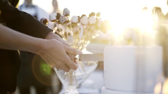 Groom and bride cuting first place of cake during wedding event. Close-up hands together holding knife and making first cut on white wedding cake in sun shining