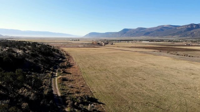 Aerial-Descending over rural mountain valley with farm fields and sagebrush and cedar tree covered hill separated by a winding dirt road.