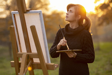 young woman artist drawing a picture on an easel in nature at sunset in autumn,concept of creativity and a hobby