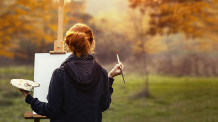 girl drawing a picture on an easel in nature, young woman with paint brush among autumn trees, a concept of creativity and a hobby - obrazy, fototapety, plakaty