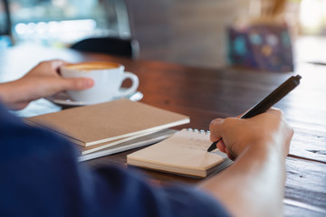 Closeup image of a woman holding and drinking hot coffee while writing on notebook on wooden table in cafe