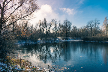 Winter landscape - forest lake with new thin ice. Trees are reflected in the water covered with new ice.
