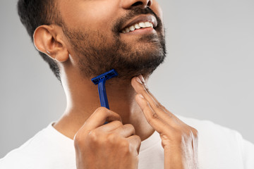 grooming and people concept - close up of young indian man shaving beard with manual razor blade over grey background