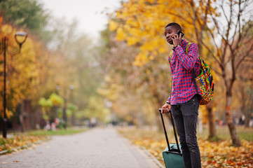 African american man in checkered shirt, with suitcase and backpack. Black man traveler at autumn park speaking on mobile phone.