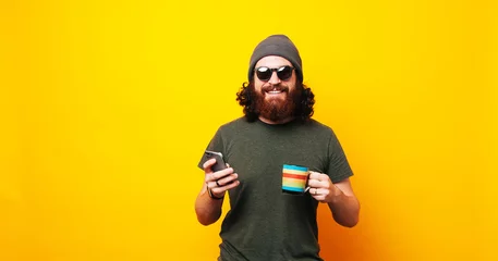 Fotobehang Happy bearded hipster man using phone and holding cup of coffee © Vulp