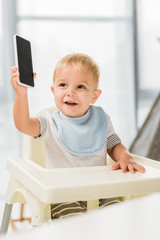 cute smiling toddler holding smartphone in hand and sitting in baby chair