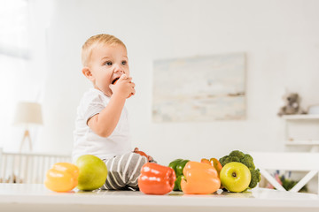Fototapeta premium cute toddler eating and sitting on table surrounded by fruit and vegetables
