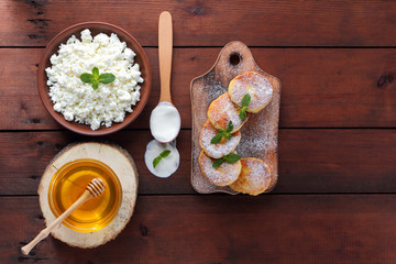 Cottage cheese pancakes sprinkled with powdered sugar. Cheesecakes on wooden board. Cottage cheese in clay bowl and sour cream in wooden spoon. Honey and dairy products. Copy space