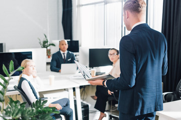 back view of young businessman holding laptop and talking with colleagues in office