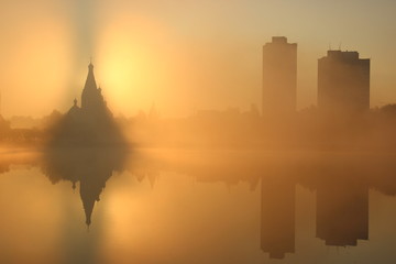 Fototapeta na wymiar Sunrise near the church, city background. Foggy morning. the mist over the water. skyscrapers around the old building