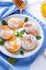 Cottage cheese pancakes sprinkled powdered sugar and fresh mint. Cheesecakes on white plate. Sour cream in wooden spoon. Honey and dairy products on white blue background. Healthy food