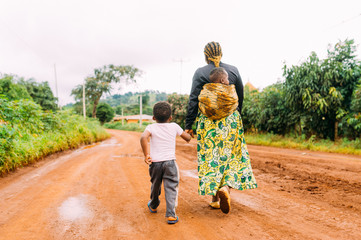 african mother and two children walk alone in red clay road in  village as a family in traditional...