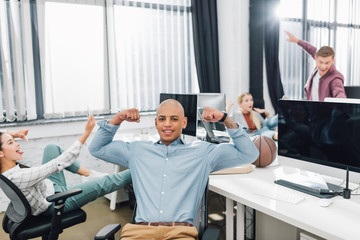 Fototapeta na wymiar happy young african american man showing biceps and smiling at camera while colleagues having fun behind in office