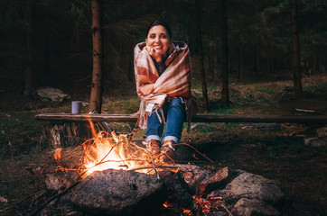 Pretty smiling young woman sits near the campfire in twilight autumn forest - Powered by Adobe