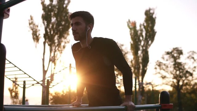Fit man doing triceps dips on parallel bars at park exercising outdoors in the morning while listening to the music in earphones. Sun shines on the background