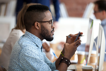 African American male employee in glasses busy with smartphone texting message to friend, serious...