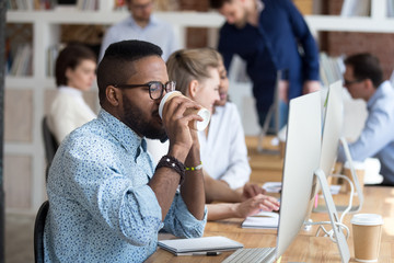 African American employee drinking coffee and talk on phone while working at computer in shared...