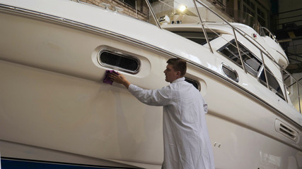 Young man (guy) specialist in white work coat polishes the yacht in the garage. Concept of: Yacht...