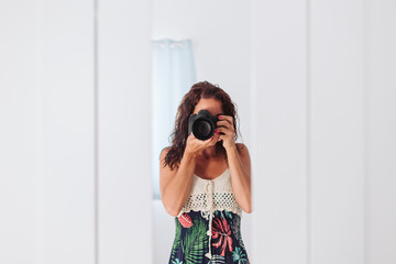 young woman taking a self portrait in the mirror at home. White walls and background. Reflex camera covering her face. Lifestyle