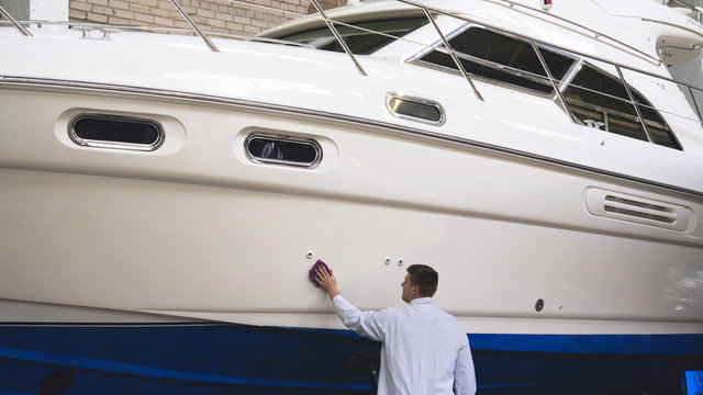 Young man (guy) specialist in white work coat polishes the yacht in the garage. Concept of: Yacht preparation, Cleaning, Polishing, Master, Professional, After painting, Using chemistry.