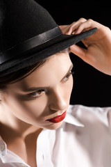 stylish pensive model with red lips in white shirt and black hat posing isolated on black