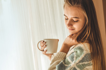Cozy home. Woman with cup of hot drink  by the window. Looking at window and drink tea. Good morning with tea. Young girl relaxing