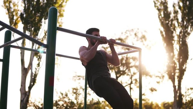 Bearded fitness man working out his arm muscles on outdoor gym doing chin-ups pull-ups as part of a crossfit workout work. Lifting up high under the bar