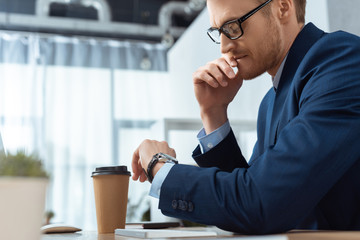 serious businessman in eyeglasses checking watch at table with paper coffee cup in office