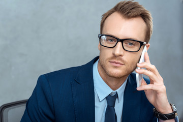 portrait of handsome businessman in eyeglasses talking on smartphone and looking at camera in office