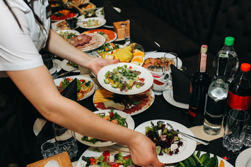 Waitress is holding fresh salad plates in her hand. Woman sets the table at the restaurant. Cafe service for birthday or wedding celebration. Different dishes on the served table.