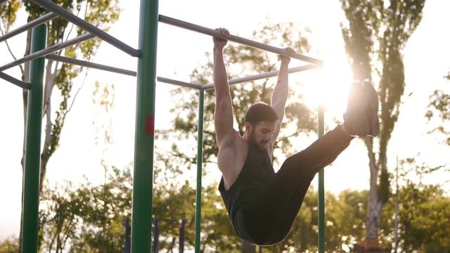 Fitness, sport, training and people concept - strong bearded man doing legs exercises while hanging on the horizontal bar in the park. Abdominal and legs exercise in outdoors gym. Slow motion