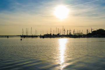 Lovely view of the sun setting behind the harbour leaving a silhouette of wind turbines and sailing boats with their mast on Fehmarn island in Germany. The sunlight is shimmering on the water surface.