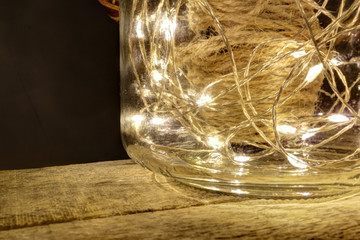 A close view of a still life composition with a glass jar with a leather cover filled with a hemp rope and some bright shining leds on a rought wood